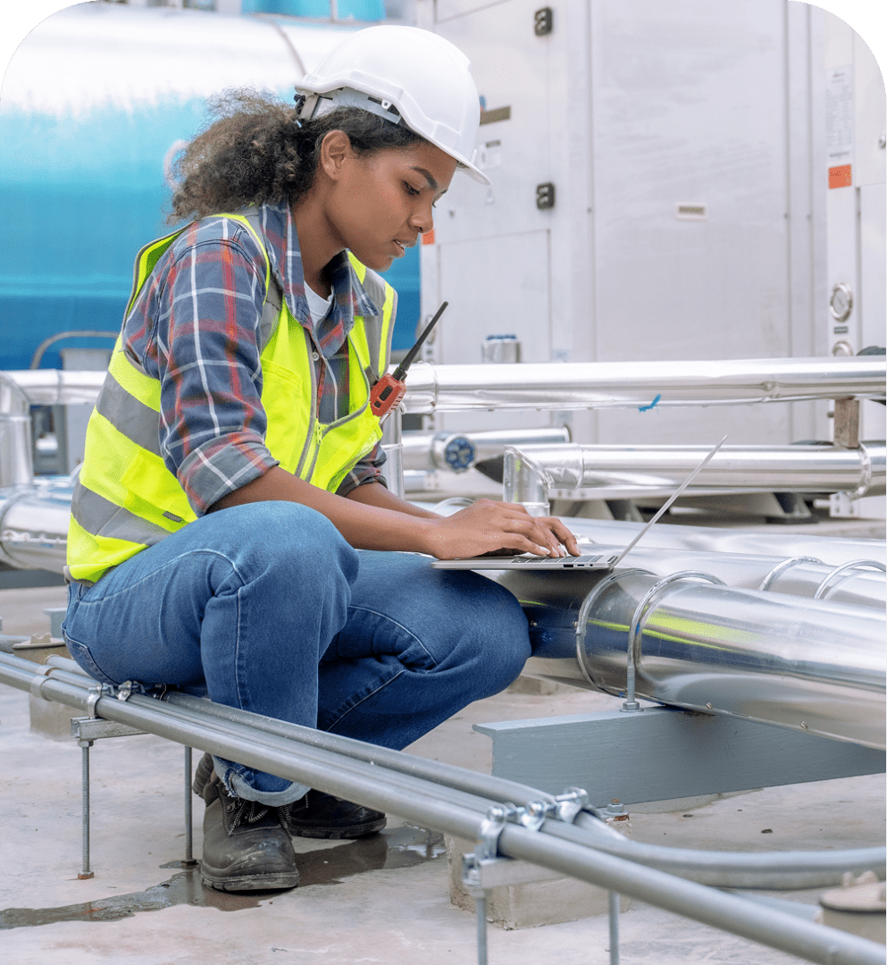 GettyImages-1414224771 1 A woman wearing a hard hat and safety vest crouches by industrial pipes, working on a laptop inside a facility.